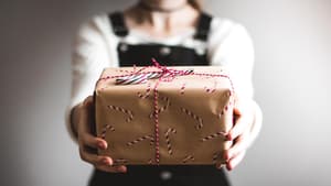Frau im schwarzen Latzkleid hält ein verpacktes Geschenk in die Kamera