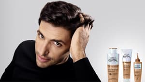 Ein Mann streicht sich mit der linken Hand durch seine dunklen, kurzen Haare und neben ihm stehen Fiber Booster Anti-Haarverlust Shampoo, Spülung und Serum von L'Oréal Paris.