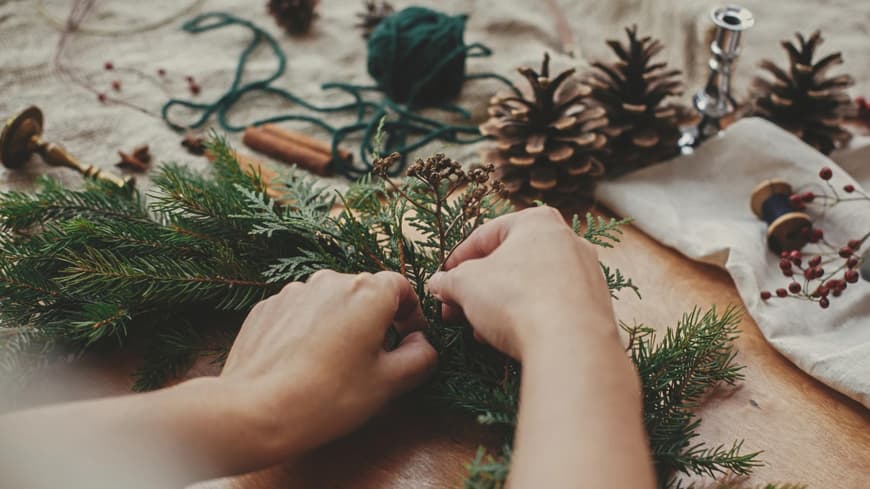 Hände mit Kräutern und Tannenzweigen, Tannenzapfen, Faden, Beeren, Zimt auf Holztisch. Weihnachtskranz-Workshop. Authentische stilvolle Stillleben