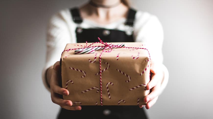 Frau im schwarzen Latzkleid hält ein verpacktes Geschenk in die Kamera