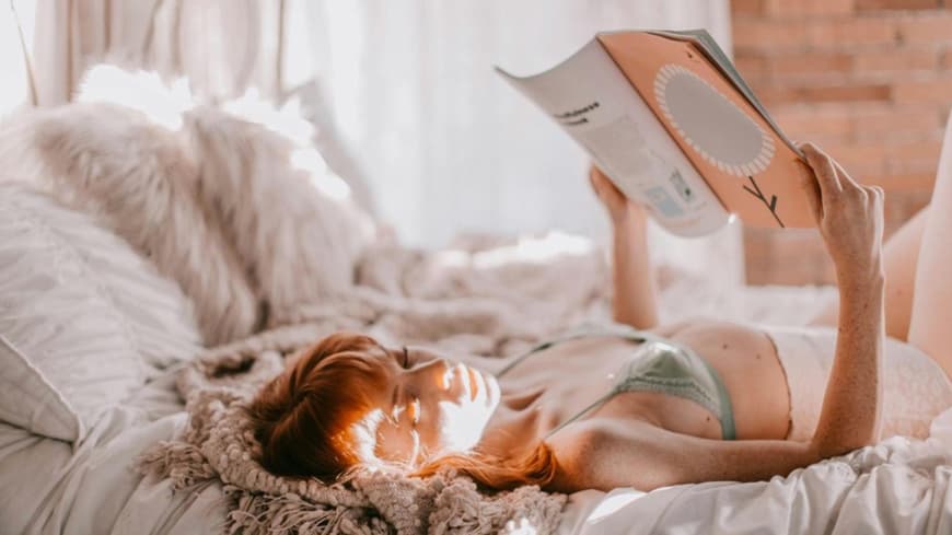 Eine junge Frau liegt in hochwertiger Wäsche in einem Bett und liest ein Magazin
