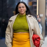 Eine curvy Frau trägt einen gelben Rock, einen Trenchcoat und einen grünen Pullover