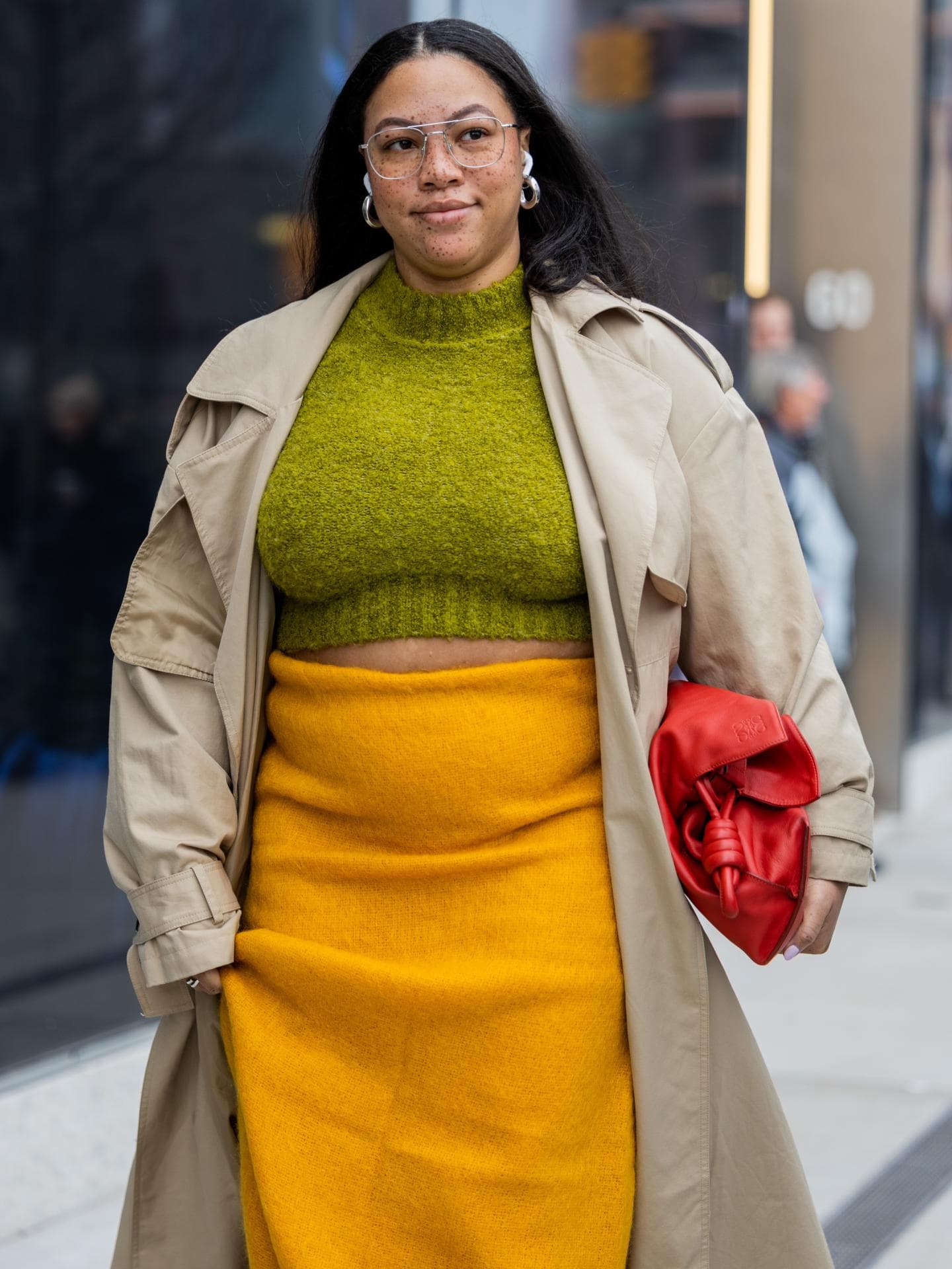 Eine curvy Frau trägt einen gelben Rock, einen Trenchcoat und einen grünen Pullover