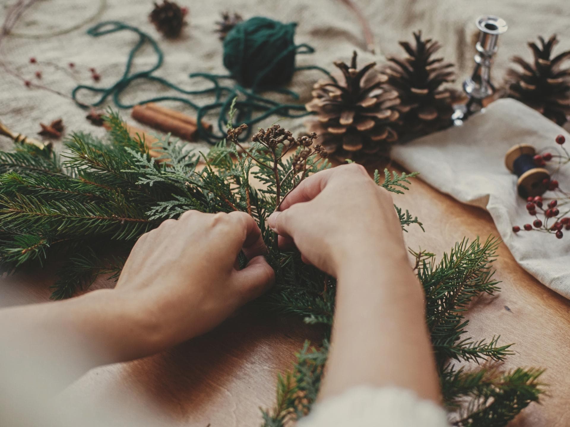 Hände mit Kräutern und Tannenzweigen, Tannenzapfen, Faden, Beeren, Zimt auf Holztisch. Weihnachtskranz-Workshop. Authentische stilvolle Stillleben Hände mit Kräutern und Tannenzweigen, Tannenzapfen, Faden, Beeren, Zimt auf Holztisch. Weihnachtskranz-Workshop. Authentische stilvolle Stillleben