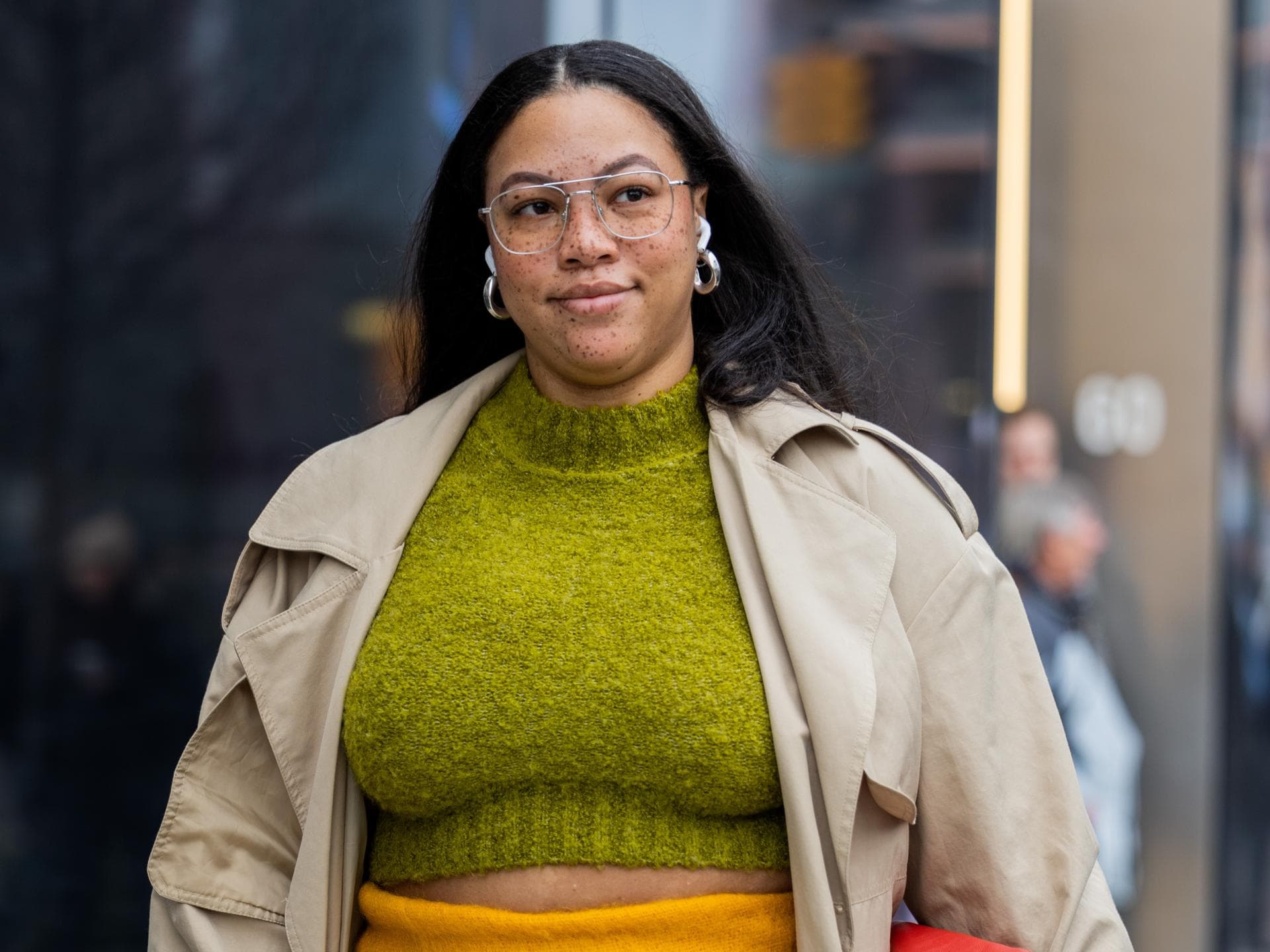 Eine curvy Frau trägt einen gelben Rock, einen Trenchcoat und einen grünen Pullover