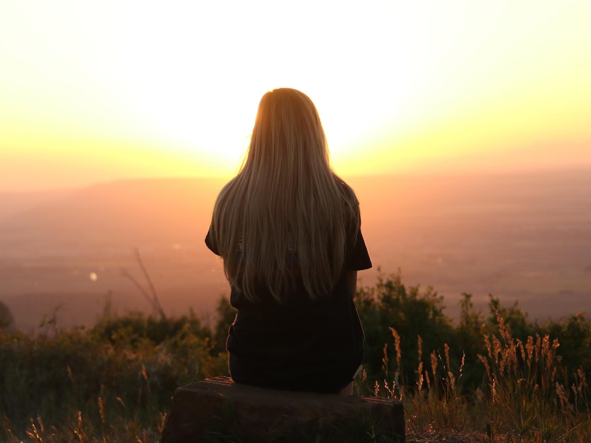 Frau sitzt auf einem Hügel und blickt in den Sonnenuntergang