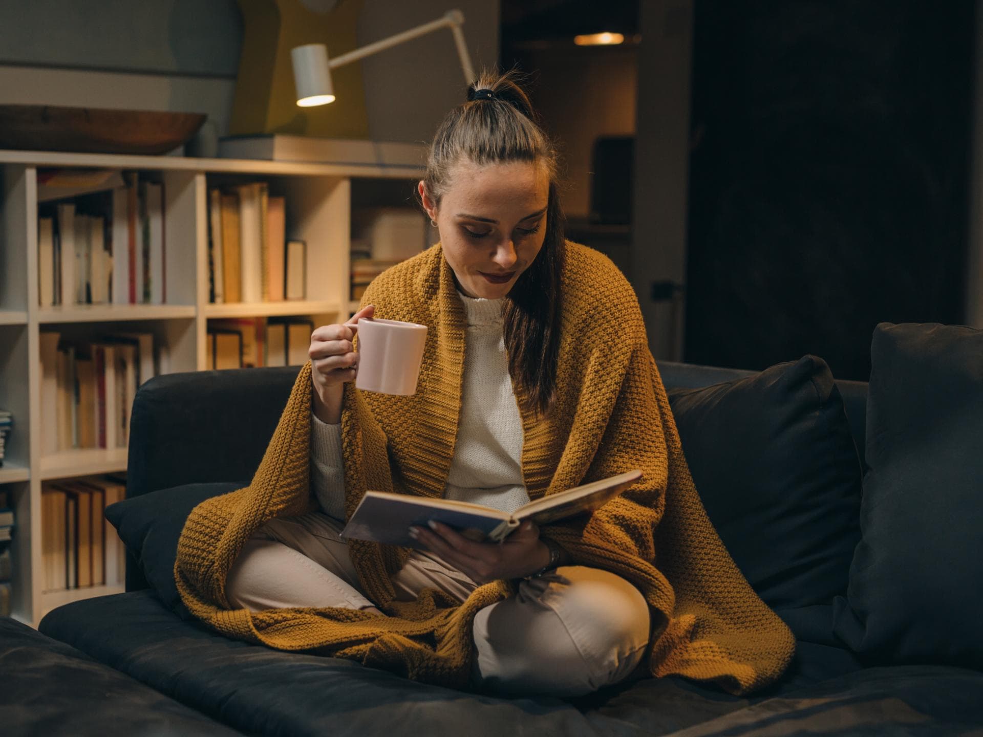 Frau mit einer gelben Decke liest ein Buch auf der Couch