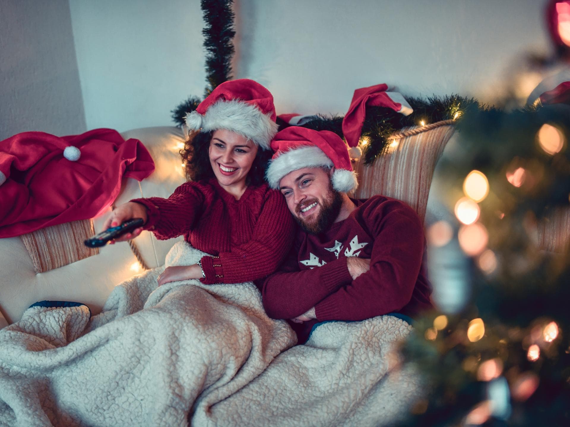Paar mit Weihnachtsmützen sitzt unter einer Decke auf der Couch