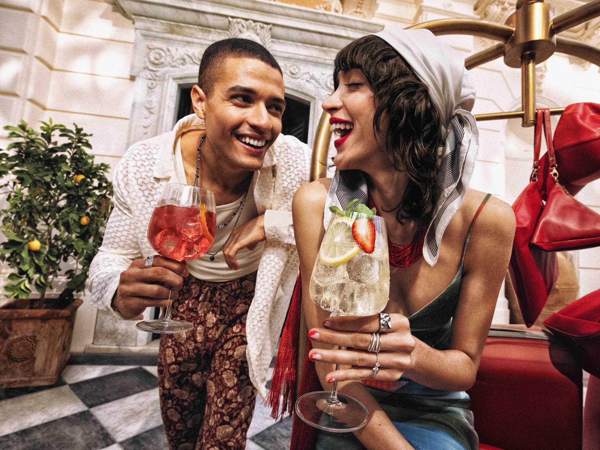 Sommerlicher Aperitivo mit Cocktails in stilvoller italienischer Atmosphäre Ein junges Paar lacht gemeinsam bei einem sommerlichen Aperitivo und hält fruchtige Cocktails in einer eleganten italienischen Hotelkulisse.