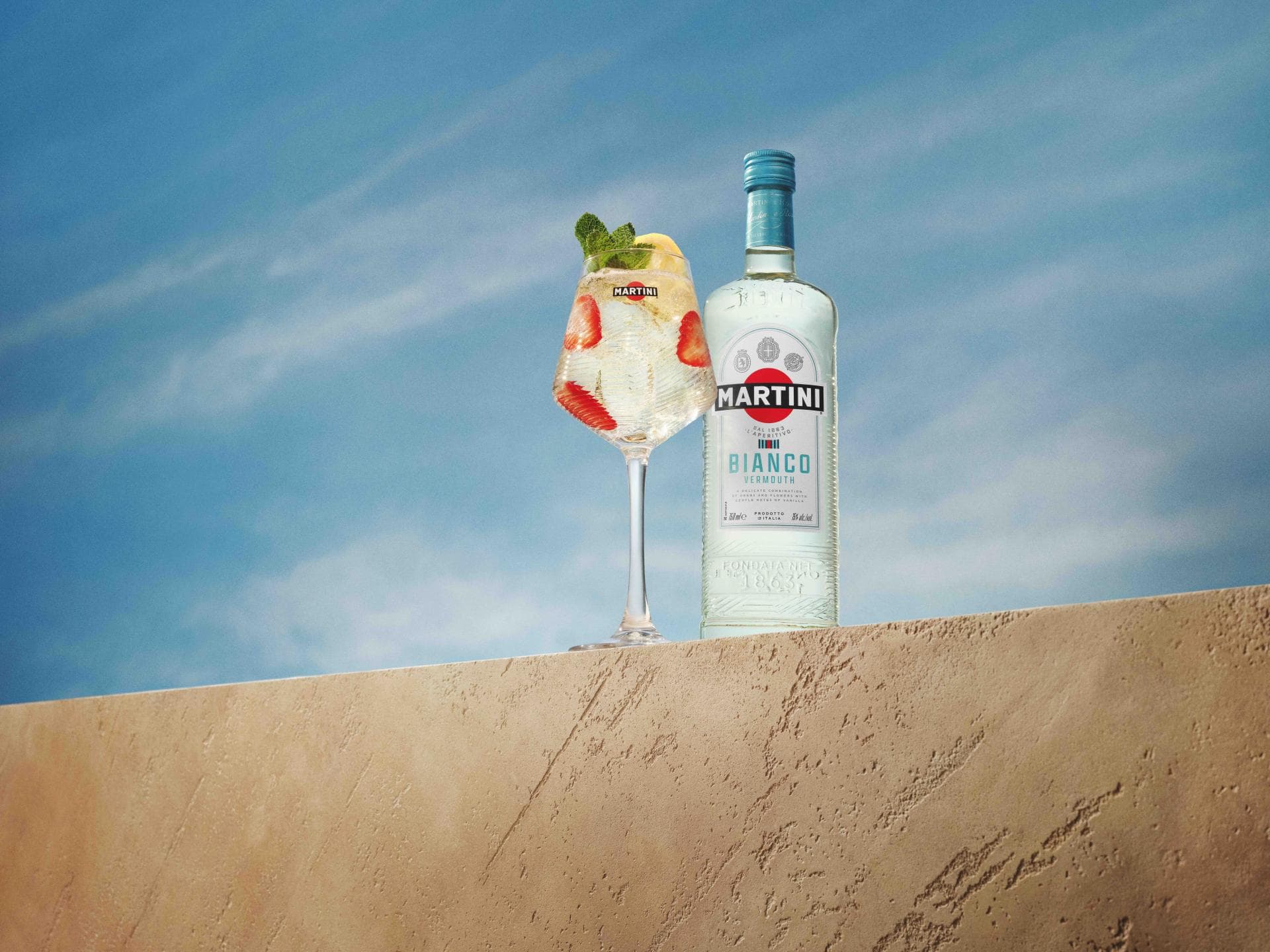 Martini Bianco Cocktail mit Erdbeeren und sommerlichem Aperitivo-Flair Eine Flasche Martini Bianco und ein sommerlicher Cocktail mit Erdbeeren und Minze stehen vor blauem Himmel auf einer hellen Steinmauer.